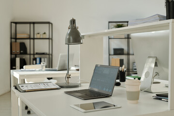 Financial graphic data on screen of laptop standing on desk among paper documents, lamp, tablet and cup of coffee in spacious office
