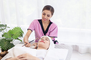 Thai professional  female masseuse therapist in uniform standing using brush brushing essential oil cream on customer face while laying down closed eyes relax on massage bed