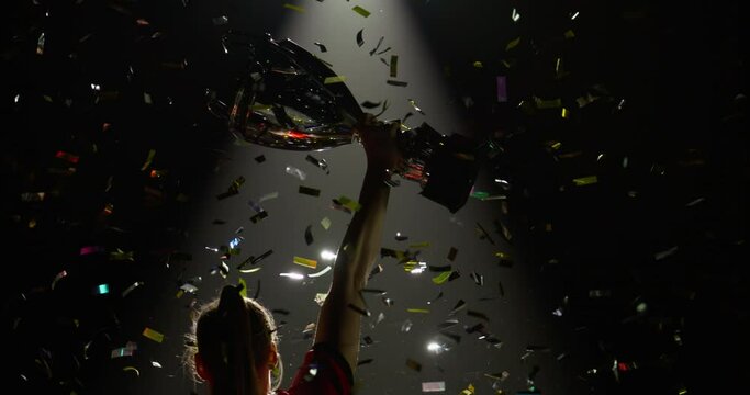 Silhouette of Caucasian woman female soccer football player raising a trophy above head against bright light and falling confetti. Super slow motion, shot on RED cinema camera