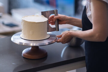 Housewife in black apron processing white mastic on organic lactose free cake with pastry scraper standing at table woman using kitchen utensils to make delicious confectionery at home