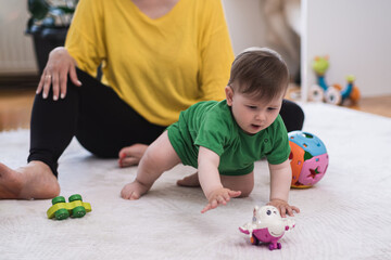 Cute little boy crawling on soft white rug near mother taking toy with hand caring woman watching...