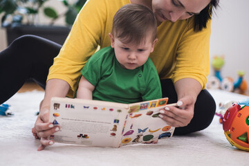 Caring mother showing focused little son book with colorful pictures of living creatures sitting on...