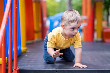 Curious preschooler with Down syndrome sitting on colorful climber at modern playground in residential complex blond kid enjoying outdoor activities in city - obrazy, fototapety, plakaty