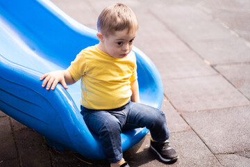 Cute little boy with Down syndrome sitting on blue slide on modern playground for kids sweet blond...