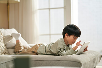 little asian boy lying on front on family couch playing computer game using digital tablet