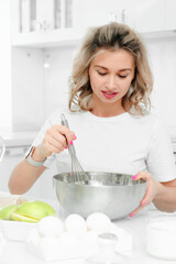 Young caucasian woman is preparing dinner in the kitchen at the table. A blonde woman in a white T-shirt stirs the dough with a whisk in a metal plate. Portrait at home.