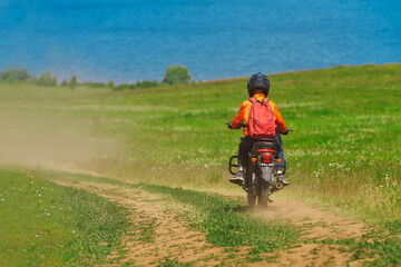Obraz premium A teenager in a protective helmet on a motorcycle on a country road at the edge of a body of water. Selective Focus.
