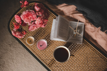 Open paper book with cup of coffee and rose flowers on rattan table in bedroom closeup. Top view. Cozy hygge atmosphere.