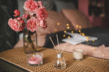Obraz na płótnie Canvas Burning candles with liquid home perfume in glass bottle on rattan table over bed indoor close up over glow lights. Cozy hygge atmosphere. Aromatherapy.