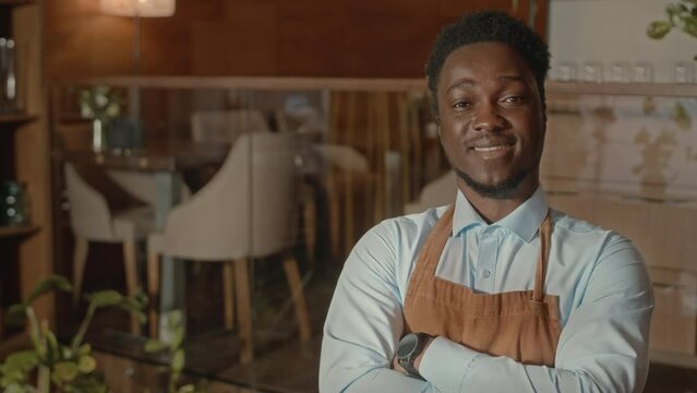 Tilt up portrait of young confident African American waiter posing for camera with hands folded working in posh expensive restaurant