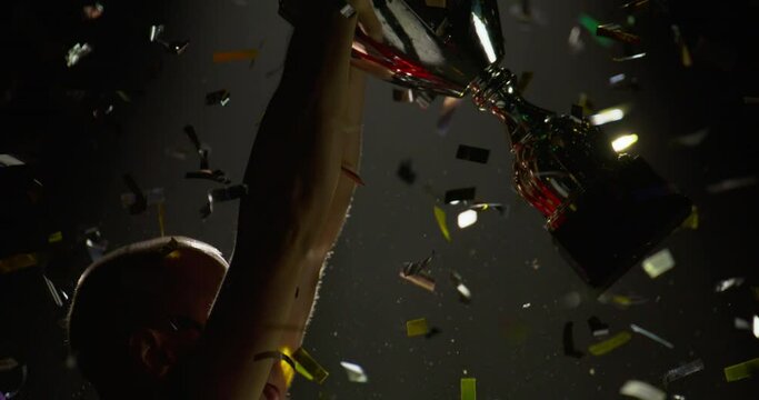 Silhouette of Caucasian male soccer football player raising a trophy above head against bright light and falling confetti. Super slow motion, shot on RED cinema camera