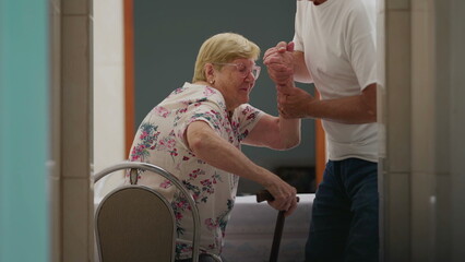 Assisting Elderly woman at home, candid authentic scene of adult son helping senior mother to get up from chair and walk through corridor