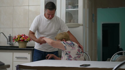 Caretaking moment of middle-aged son helping elderly mother get up from chair at home, real life...