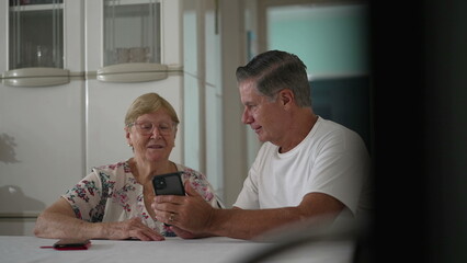 Candid man holding phone at home kitchen showing content online to senior woman in her 80s. Domestic lifestyle scene of old age concept with modern technology