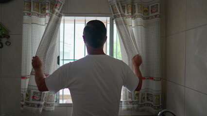 Back of person opening kitchen curtain in the morning, man starting the day and gazing out at view daylight