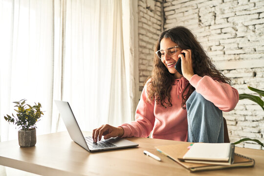 A young woman, focused and productive, working at her home desk with a laptop, multitasking while on a phone call.