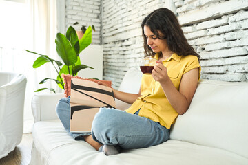 A young woman, seated on the sofa, savoring a cup of tea and engrossed in a magazine, enjoying a...