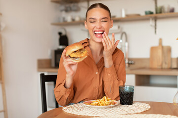 Excited young lady eating burger and laughing, eating a harmful food, sitting at table in kitchen...