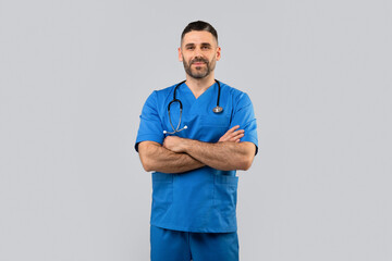 Middle aged man doctor in uniform with crossed arms on chest looking at camera on light gray background, free space