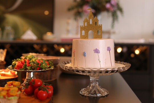 Mesa montada para evento social com comidas e bolo de anivers&aacute;rio em tema de castelo de princesa. O bolo, decorado com um castelo dourado brilhante e flores, &eacute; rosa.
