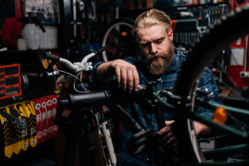 Naklejka na ściany i meble Handsome bearded cycling mechanic male repairing and fixing mountain bicycle standing on bike rack working in bike repair shop with dark interior. Concept of maintenance of bicycle transport.
