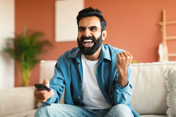 Joyful indian man shaking fists and shouting watching TV indoor