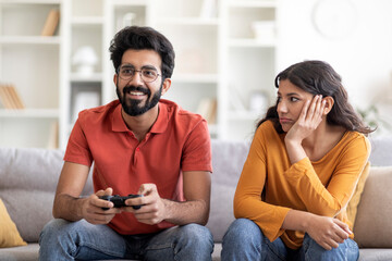 Bored young indian woman looking at boyfriend playing video games at home