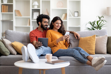 Happy Young Indian Couple Relaxing At Home With Smartphone And Digital Tablet