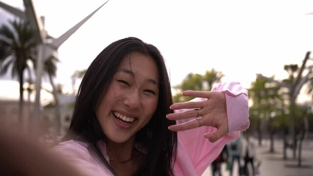 Happy Chinese young woman in a video call or live streaming her summer trip. Female student taking a selfie outdoors waving looking at camera. Point of view.