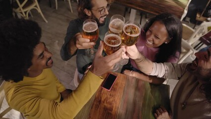 Group of multirracial young people toasting with a pint of beer and celebrating happy hours in a bar indoors. Friends drink alcohol together having fun and smiling. Cheers of gathering latin students
