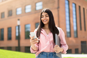 Fototapeta premium Social networks for study. Happy lady student with phone and laptop in hand, ready for lesson in university, outdoors