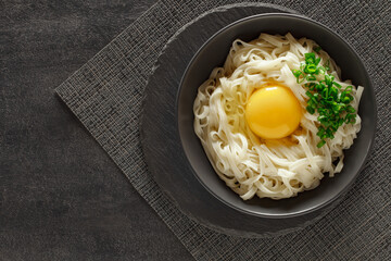 Udon noodles cooked with raw egg and green onion in a gray bowl, on slate stone plate round, dark background, top view