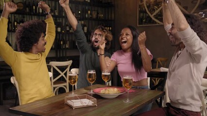Interracial group of friends fans of football celebrating a goal of victory in a bar indoors watching tv. People supporting the competition winning team with excitement and encouragement.