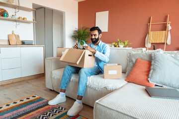 Cheerful indian man opening carton parcel, receiving purchased headphones indoors