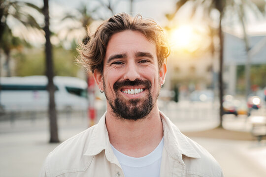 Close up portrait of handsome bearded guy with perfect white teeth smiling and looking at camera standing outdoors. Front view of young adult man with friendly and positive expression. Happy male