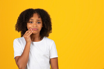 Sad curly teenager black schoolgirl in white t-shirt presses hand to cheek, suffering from toothache