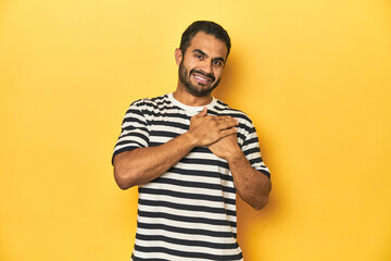 Casual young Latino man against a vibrant yellow studio background, has friendly expression, pressing palm to chest. Love concept.