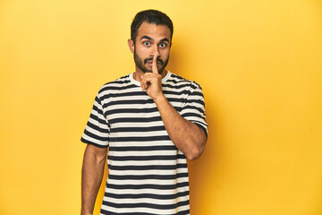 Casual young Latino man against a vibrant yellow studio background, keeping a secret or asking for silence.