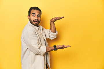 Casual young Latino man against a vibrant yellow studio background, shocked and amazed holding a copy space between hands.