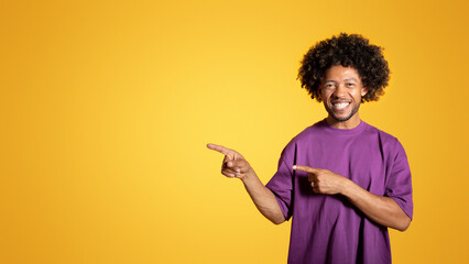 Glad black adult curly guy in purple t-shirt points fingers at empty space, isolated on orange background