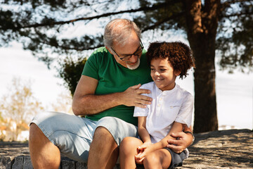 Fototapeta premium Glad senior man with beard and black little boy hugging, have fun, relaxing in park, enjoy weekend
