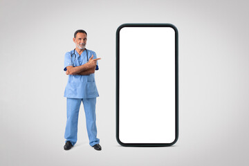 Happy caucasian mature man doctor in uniform point on huge phone with blank screen