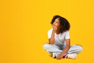 Happy curly teenager black schoolgirl in white t-shirt sits on floor, looks at free space, isolated...