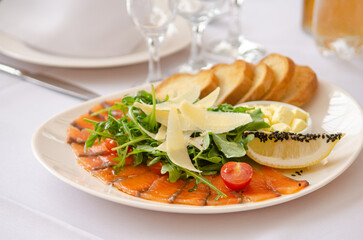Restaurant meal, sliced red fish, bread, greens, lemon and cheese on a white plate, among other dishes on a white tablecloth