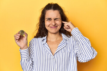 Woman holding a physical Bitcoin coin, covering ears with hands.