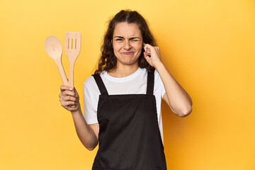 Woman with apron, wooden cooking utensils, yellow, covering ears with hands.