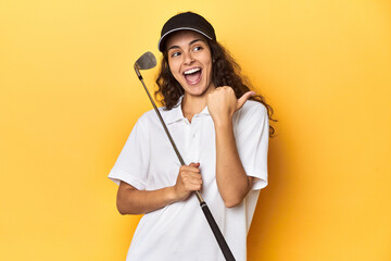 Golfer woman with cap, golf polo, yellow studio, points with thumb finger away, laughing and carefree.