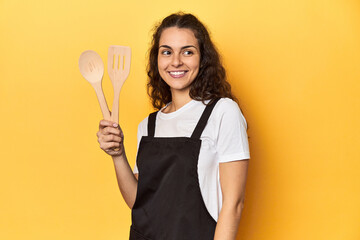 Woman with apron, wooden cooking utensils, yellow, looks aside smiling, cheerful and pleasant.