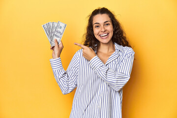 Delighted woman holding dollars, bright yellow backdrop.