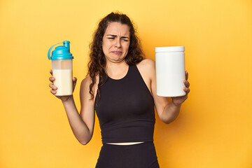 Young Caucasian woman with protein jar and shake, in sportswear, yellow background.